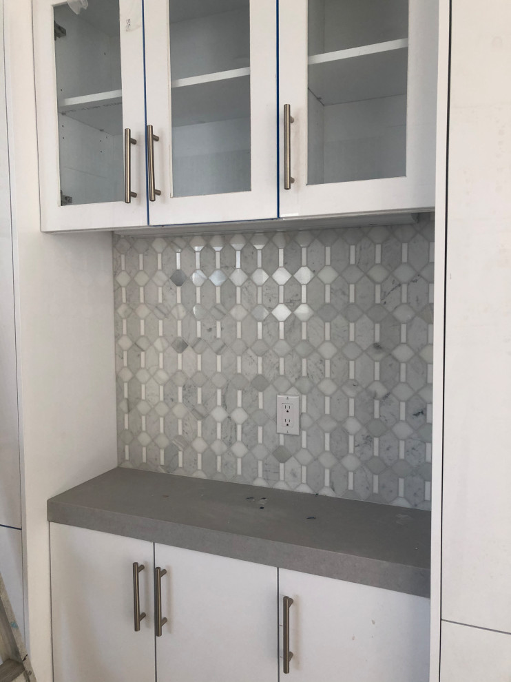 This is an example of a mid-sized transitional single-wall home bar in Los Angeles with flat-panel cabinets, white cabinets, quartz benchtops, grey splashback, marble splashback, light hardwood floors and grey benchtop.