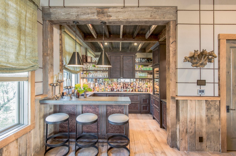 This is an example of a large country u-shaped seated home bar in Austin with an undermount sink, raised-panel cabinets, brown cabinets, mirror splashback, light hardwood floors and grey benchtop.