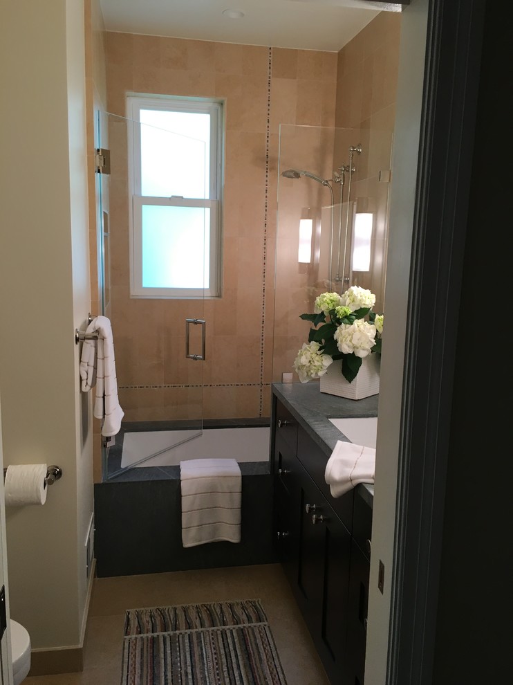 This is an example of a small transitional kids bathroom in San Francisco with recessed-panel cabinets, dark wood cabinets, an undermount tub, a shower/bathtub combo, yellow tile, limestone, beige walls, mosaic tile floors, an undermount sink, soapstone benchtops, multi-coloured floor and a hinged shower door.