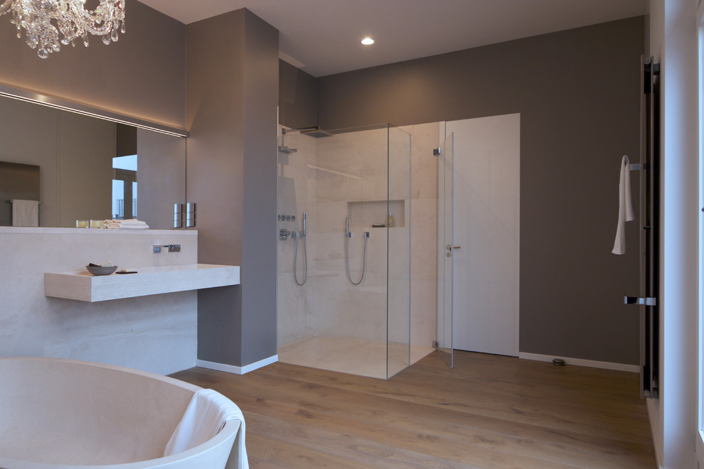 Inspiration for a large contemporary master bathroom in Berlin with a freestanding tub, a curbless shower, a wall-mount toilet, beige tile, grey walls, medium hardwood floors, a wall-mount sink, brown floor and a hinged shower door.