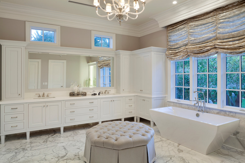 This is an example of a large traditional master bathroom in Miami with white cabinets, a freestanding tub, raised-panel cabinets, beige tile, beige walls, marble floors, an undermount sink, engineered quartz benchtops, white floor, a hinged shower door and white benchtops.