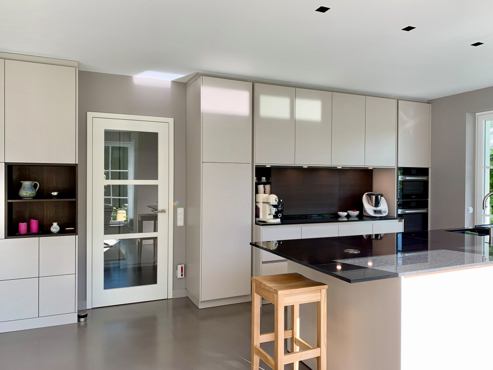 This is an example of an expansive contemporary single-wall eat-in kitchen in Hamburg with an undermount sink, flat-panel cabinets, beige cabinets, granite benchtops, black splashback, timber splashback, black appliances, concrete floors, with island, beige floor and black benchtop.