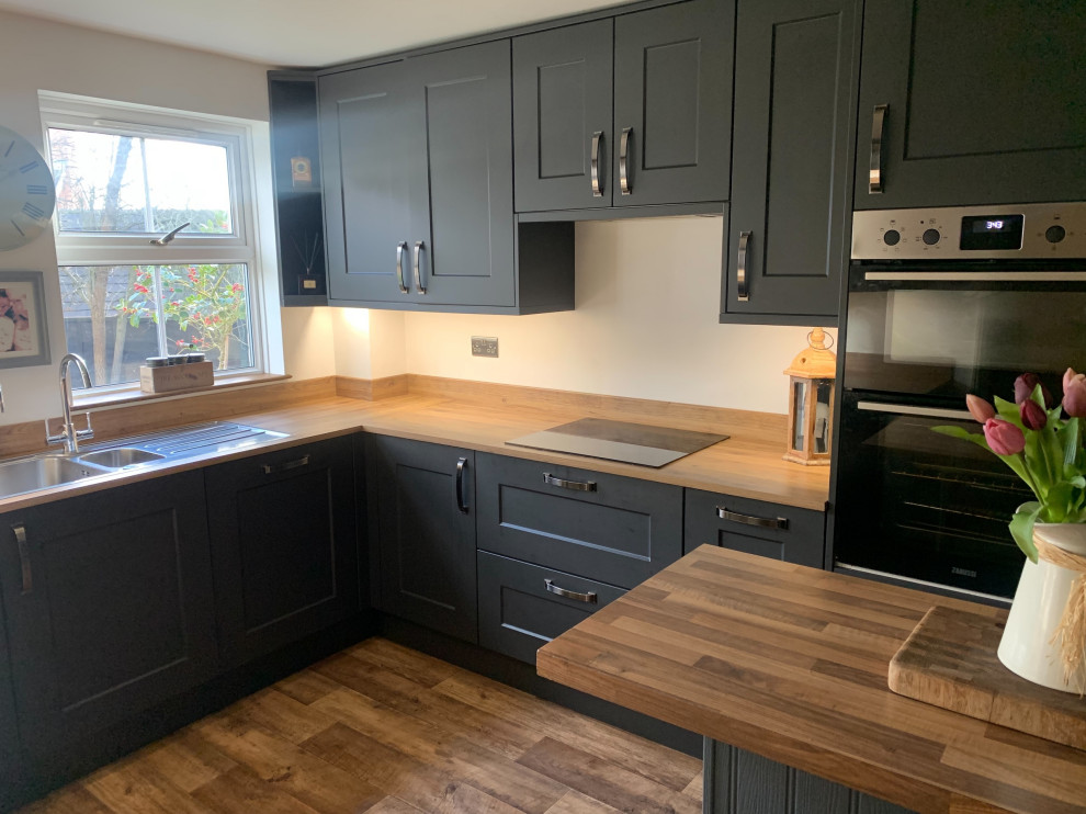 Photo of a medium sized traditional u-shaped enclosed kitchen in Surrey with a double-bowl sink, recessed-panel cabinets, black cabinets, laminate countertops, black appliances, medium hardwood flooring, an island, brown floors and brown worktops.