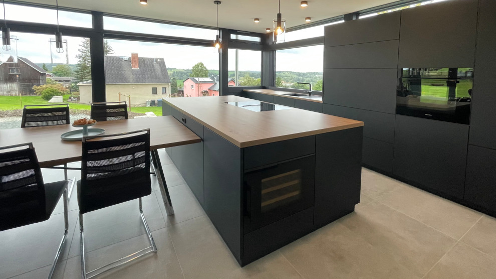 Design ideas for a large modern galley open plan kitchen in Nuremberg with a drop-in sink, flat-panel cabinets, black cabinets, wood benchtops, white splashback, black appliances, concrete floors, with island, grey floor and brown benchtop.