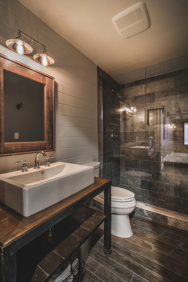 Inspiration for a mid-sized country 3/4 bathroom in Columbus with open cabinets, distressed cabinets, a one-piece toilet, gray tile, ceramic tile, grey walls, ceramic floors, wood benchtops, a vessel sink, brown benchtops, an alcove shower, brown floor and a hinged shower door.