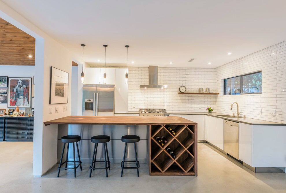 Photo of a mid-sized contemporary u-shaped eat-in kitchen in Los Angeles with an undermount sink, flat-panel cabinets, white cabinets, wood benchtops, white splashback, stainless steel appliances, concrete floors, a peninsula, grey floor, brown benchtop and subway tile splashback.
