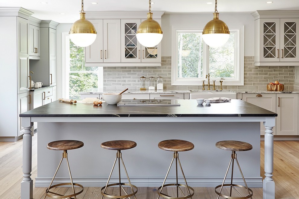 Example of a transitional light wood floor and beige floor kitchen design with a farmhouse sink, shaker cabinets, white cabinets, gray backsplash, subway tile backsplash and an island