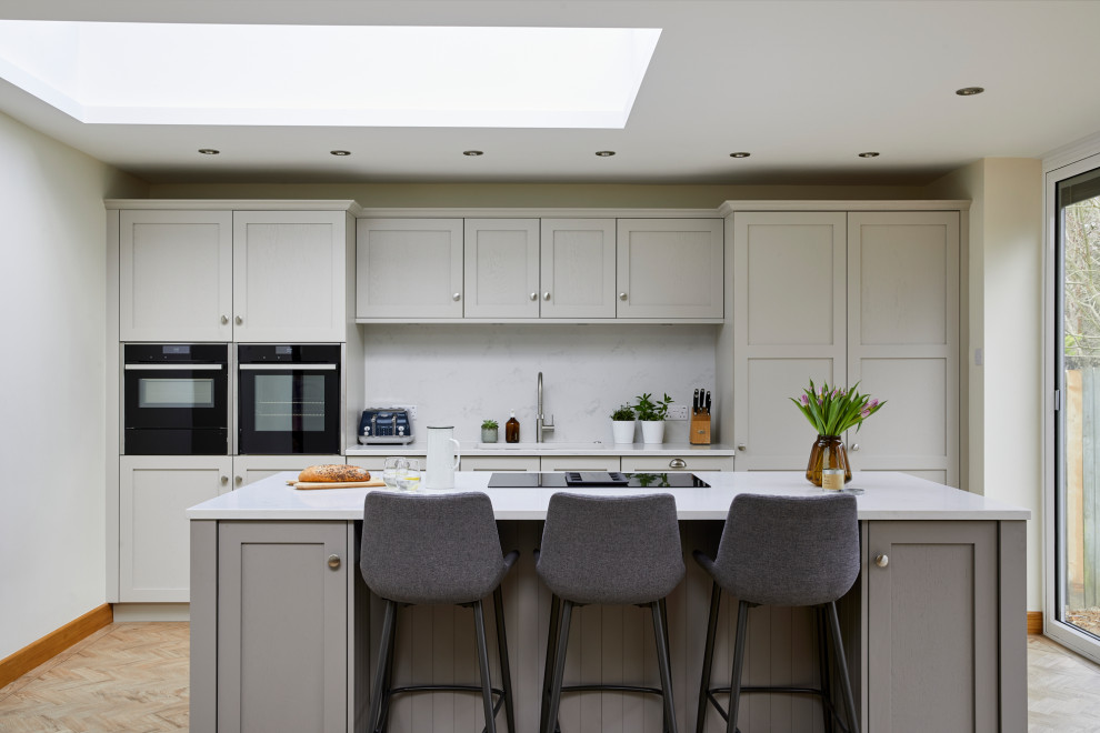 Imagen de cocina comedor lineal clásica de tamaño medio con fregadero bajoencimera, armarios estilo shaker, puertas de armario grises, encimera de cuarcita, salpicadero blanco, puertas de cuarzo sintético, electrodomésticos con paneles, suelo de madera en tonos medios, una isla, suelo marrón y encimeras blancas