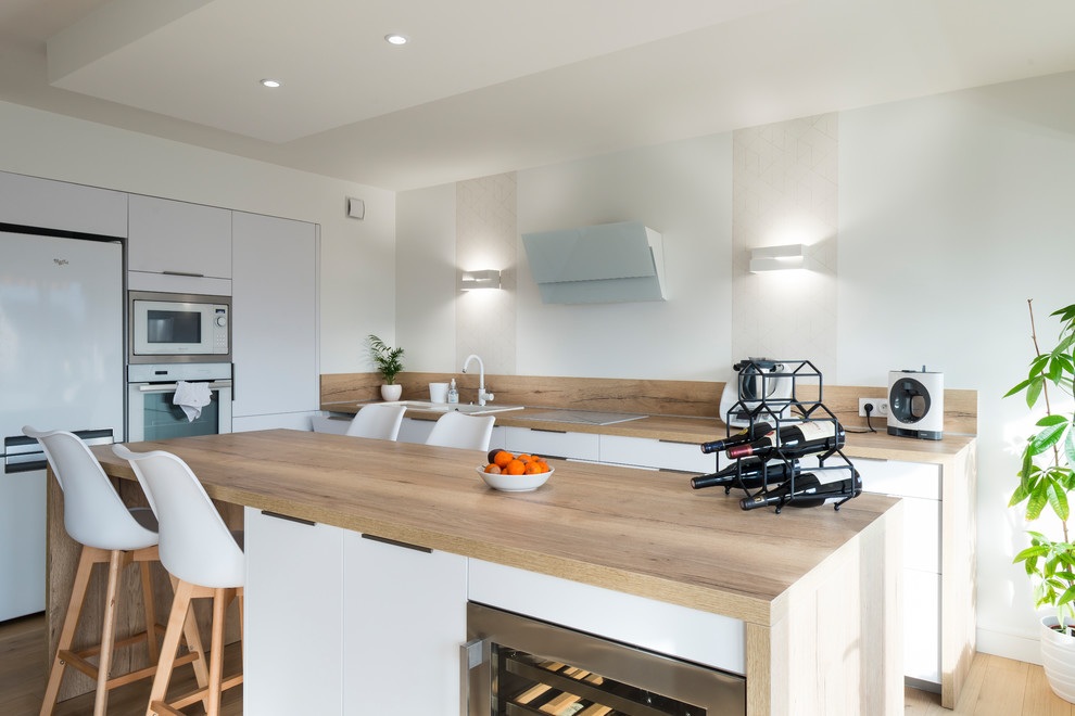 This is an example of a large scandinavian kitchen in Grenoble with white cabinets, wood benchtops, beige splashback, timber splashback, light hardwood floors, with island, beige floor, beige benchtop, a drop-in sink, flat-panel cabinets and white appliances.