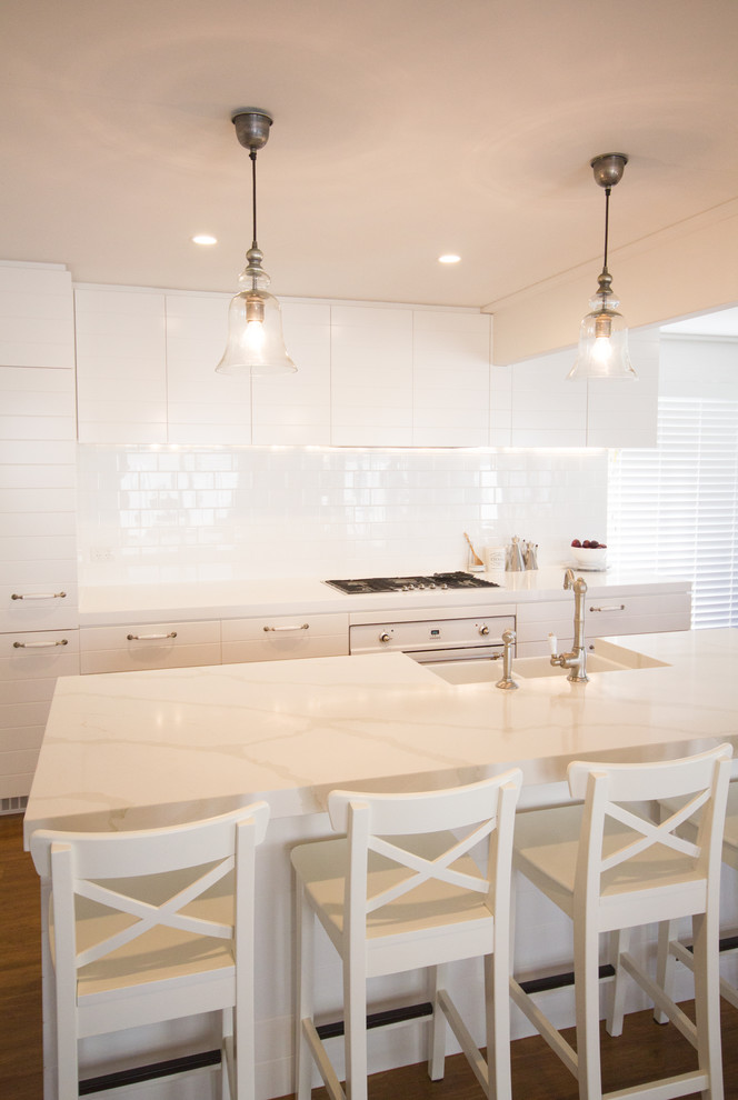 This is an example of a beach style galley eat-in kitchen in Gold Coast - Tweed with with island, white cabinets, quartz benchtops, white splashback, ceramic splashback, white appliances, a farmhouse sink and bamboo floors.