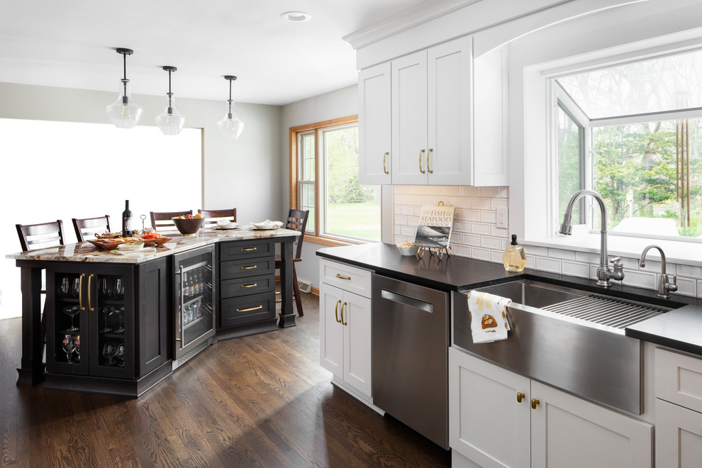 Large traditional u-shaped eat-in kitchen in Milwaukee with a farmhouse sink, recessed-panel cabinets, white cabinets, granite benchtops, white splashback, subway tile splashback, stainless steel appliances, medium hardwood floors, with island, brown floor and black benchtop.