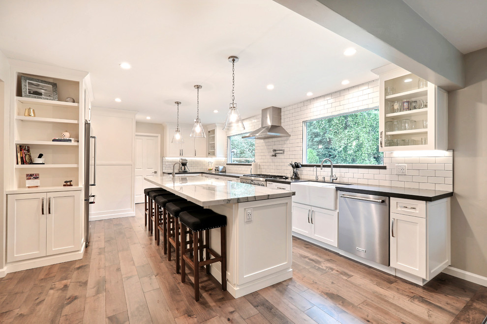 Photo of a large transitional l-shaped eat-in kitchen in San Francisco with a farmhouse sink, shaker cabinets, white cabinets, quartz benchtops, white splashback, subway tile splashback, stainless steel appliances, medium hardwood floors, with island, brown floor and black benchtop.