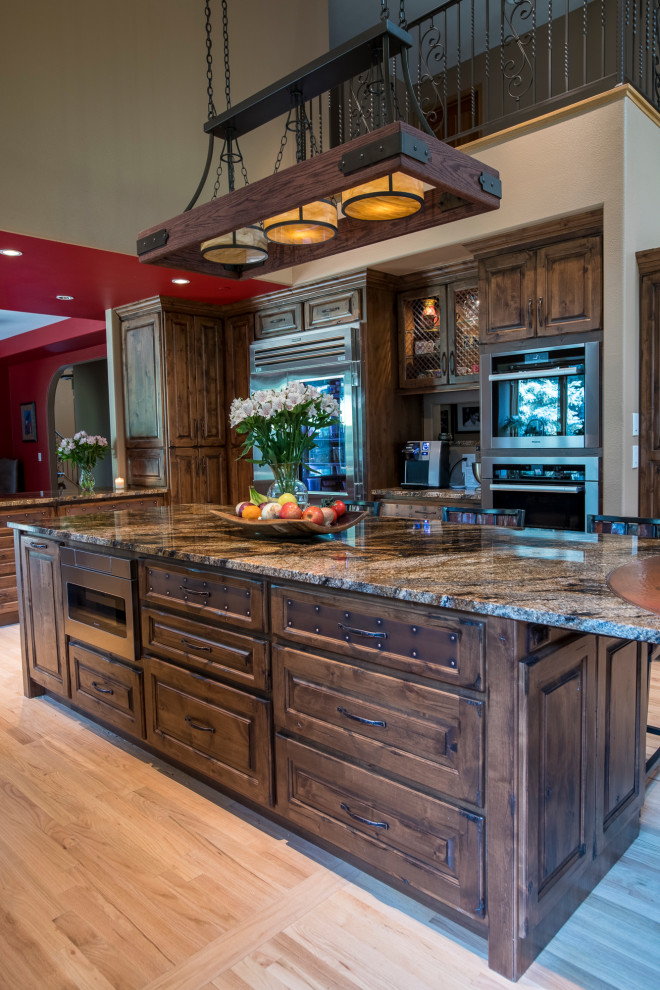Example of a large classic light wood floor, beige floor and vaulted ceiling eat-in kitchen design in Denver with an undermount sink, recessed-panel cabinets, distressed cabinets, granite countertops, beige backsplash, stone tile backsplash, stainless steel appliances, an island and brown countertops