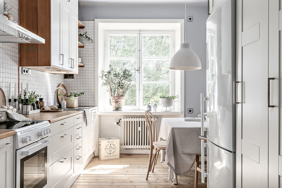 Design ideas for a mid-sized scandinavian eat-in kitchen in Stockholm with white cabinets, wood benchtops, white appliances, light hardwood floors, no island, beige floor, an undermount sink, white splashback, beige benchtop and shaker cabinets.