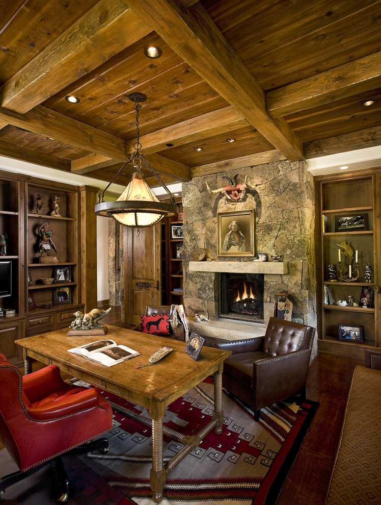 This is an example of a mid-sized country home office in Other with dark hardwood floors, a ribbon fireplace, a stone fireplace surround, a freestanding desk, brown floor and exposed beam.