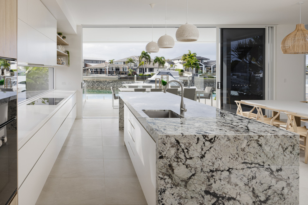Photo of a large contemporary galley open plan kitchen in Brisbane with an undermount sink, flat-panel cabinets, white cabinets, marble benchtops, glass sheet splashback, black appliances, porcelain floors, with island, beige floor and white benchtop.