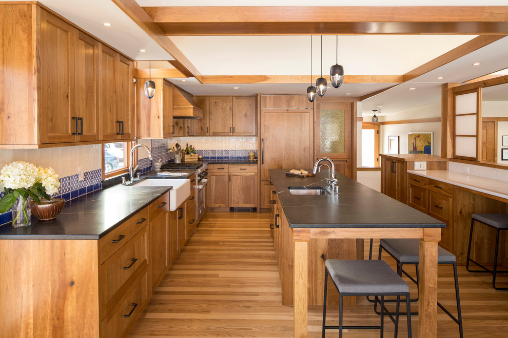Design ideas for a country l-shaped kitchen in Minneapolis with an undermount sink, shaker cabinets, medium wood cabinets, blue splashback, panelled appliances, light hardwood floors, multiple islands, beige floor and black benchtop.