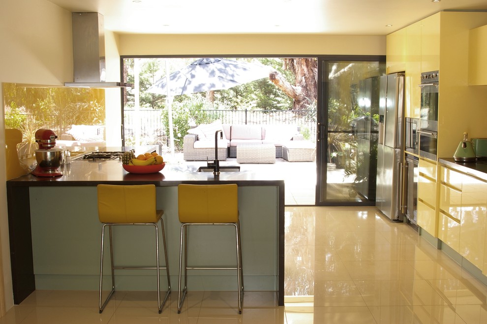 Photo of a mid-sized contemporary l-shaped open plan kitchen in Adelaide with an undermount sink, flat-panel cabinets, yellow cabinets, quartz benchtops, yellow splashback, glass sheet splashback, stainless steel appliances, porcelain floors, a peninsula and beige floor.
