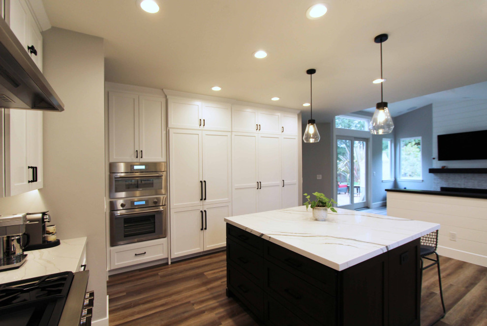 Example of a large transitional l-shaped vinyl floor and brown floor open concept kitchen design with a farmhouse sink, shaker cabinets, white cabinets, quartz countertops, white backsplash, ceramic backsplash, stainless steel appliances, an island and white countertops