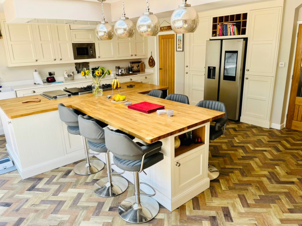 Photo of an expansive traditional kitchen in Buckinghamshire with a farmhouse sink, shaker cabinets, white cabinets, wood benchtops, white splashback, engineered quartz splashback, black appliances, vinyl floors, with island, brown floor, white benchtop and recessed.