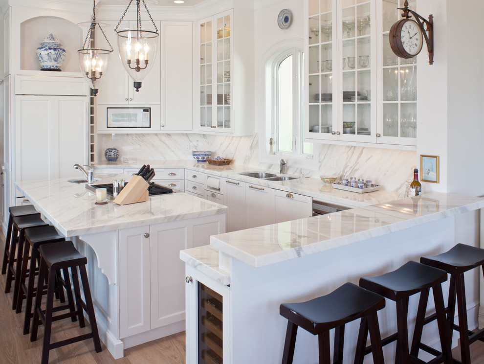 Photo of a mid-sized traditional u-shaped eat-in kitchen in Tampa with glass-front cabinets, an undermount sink, white cabinets, marble benchtops, white splashback, marble splashback, panelled appliances, light hardwood floors, with island and brown floor.