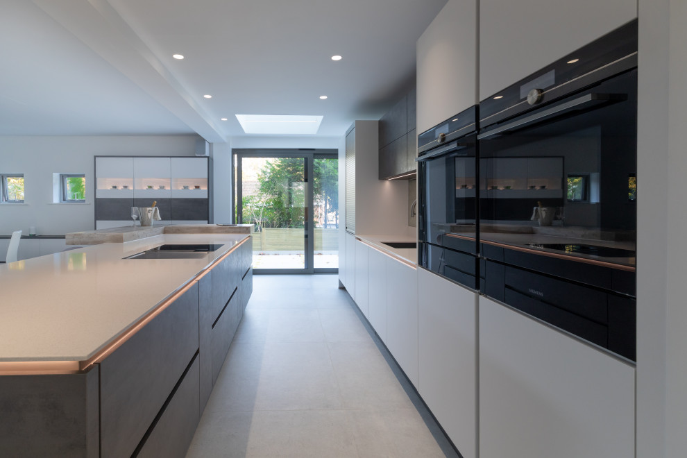 Design ideas for a large contemporary single-wall eat-in kitchen in Hampshire with a single-bowl sink, flat-panel cabinets, white cabinets, quartzite benchtops, multi-coloured splashback, mirror splashback, black appliances, ceramic floors, with island, grey floor and white benchtop.