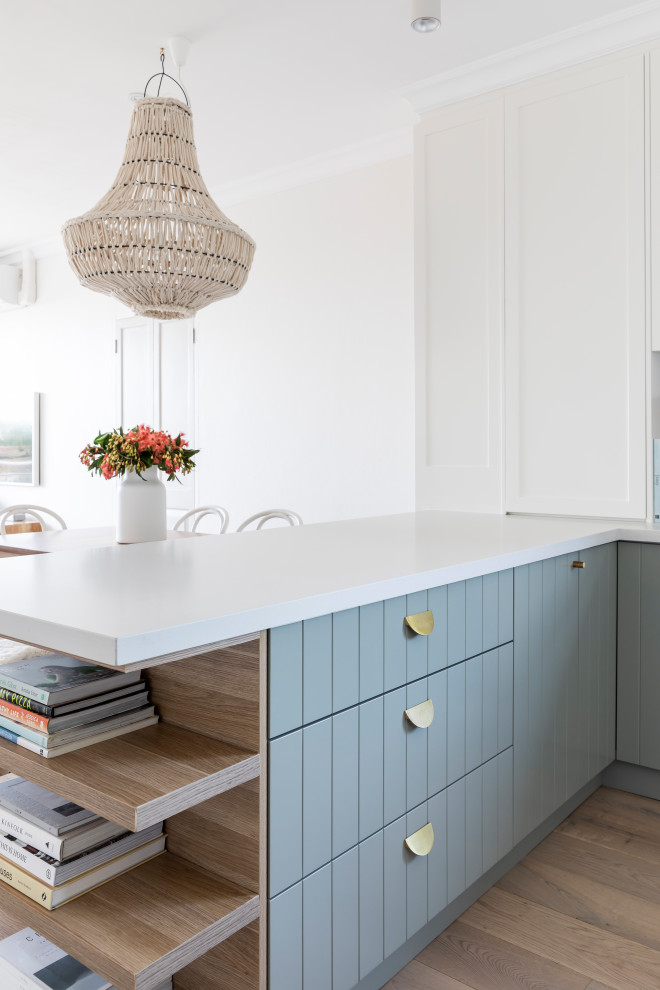 Photo of a small beach style u-shaped eat-in kitchen in Sydney with an undermount sink, shaker cabinets, white cabinets, quartz benchtops, white splashback, marble splashback, stainless steel appliances, light hardwood floors, no island, brown floor and white benchtop.