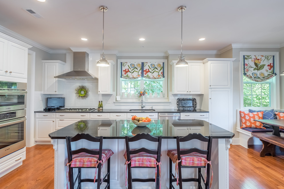 Photo of a traditional l-shaped eat-in kitchen in Philadelphia with an undermount sink, raised-panel cabinets, white cabinets, white splashback, stainless steel appliances, medium hardwood floors, with island and brown floor.