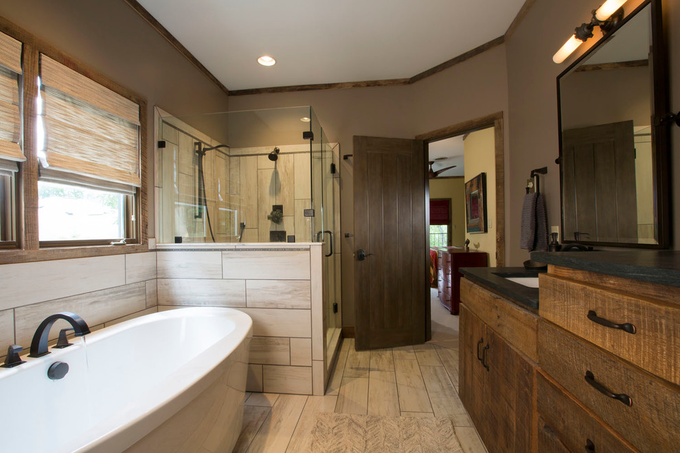 Mid-sized country master bathroom in Other with flat-panel cabinets, dark wood cabinets, a freestanding tub, a corner shower, a two-piece toilet, beige tile, brown walls, vinyl floors, an undermount sink, soapstone benchtops, beige floor and a hinged shower door.