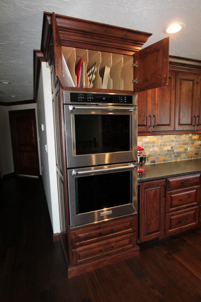 Large mountain style medium tone wood floor and brown floor kitchen photo with raised-panel cabinets, medium tone wood cabinets, granite countertops, multicolored backsplash, stone tile backsplash, paneled appliances, an island and black countertops