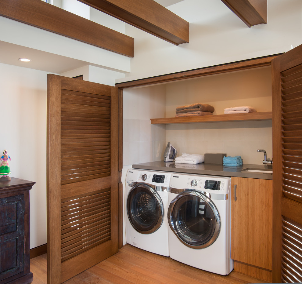 Photo of a small beach style single-wall laundry cupboard in San Diego with an undermount sink, flat-panel cabinets, solid surface benchtops, white walls, light hardwood floors, a side-by-side washer and dryer and medium wood cabinets.