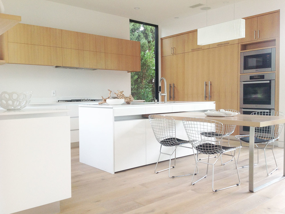 This is an example of a mid-sized contemporary u-shaped eat-in kitchen in Los Angeles with a drop-in sink, flat-panel cabinets, white cabinets, soapstone benchtops, white splashback, stone tile splashback, stainless steel appliances, light hardwood floors and with island.