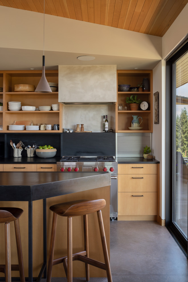 Mid-sized modern single-wall eat-in kitchen in Portland with light wood cabinets, soapstone benchtops, black splashback, stainless steel appliances, concrete floors, with island, black benchtop, wood, flat-panel cabinets, stone slab splashback and grey floor.