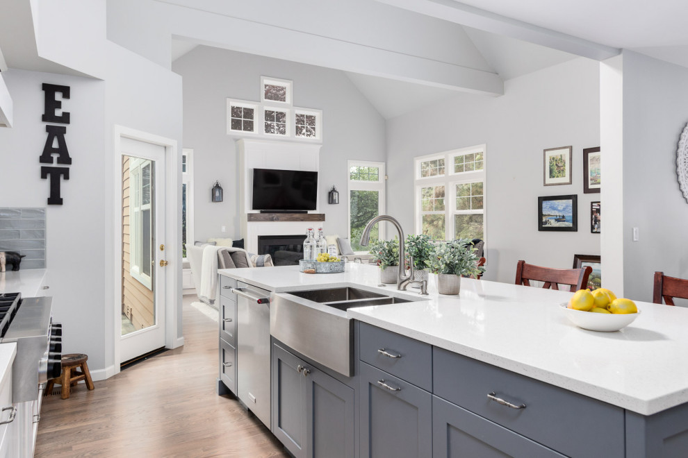 Mid-sized transitional l-shaped open plan kitchen in Seattle with a farmhouse sink, shaker cabinets, grey cabinets, quartz benchtops, grey splashback, subway tile splashback, stainless steel appliances, light hardwood floors, with island and white benchtop.