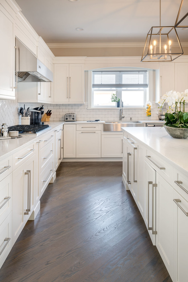 This is an example of a mid-sized transitional l-shaped separate kitchen in New York with a farmhouse sink, shaker cabinets, white cabinets, quartzite benchtops, white splashback, subway tile splashback, stainless steel appliances, medium hardwood floors, with island, brown floor and white benchtop.