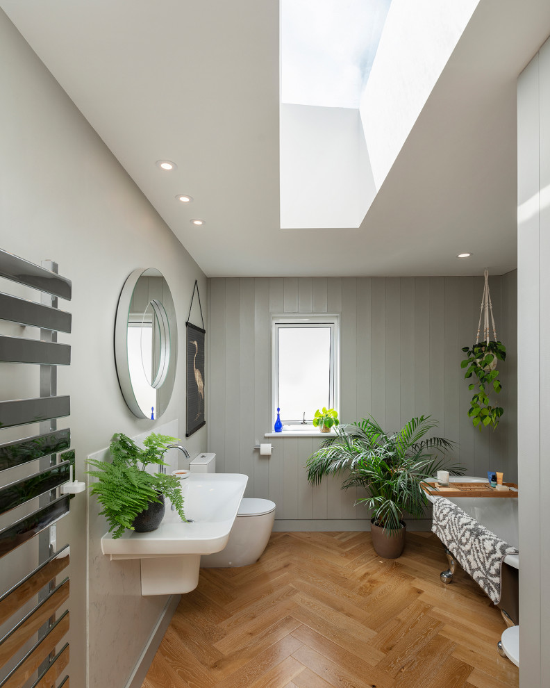 Design ideas for a mid-sized contemporary kids bathroom in Devon with a freestanding tub, a one-piece toilet, white tile, ceramic tile, grey walls, medium hardwood floors, a wall-mount sink, brown floor, a single vanity, vaulted and panelled walls.