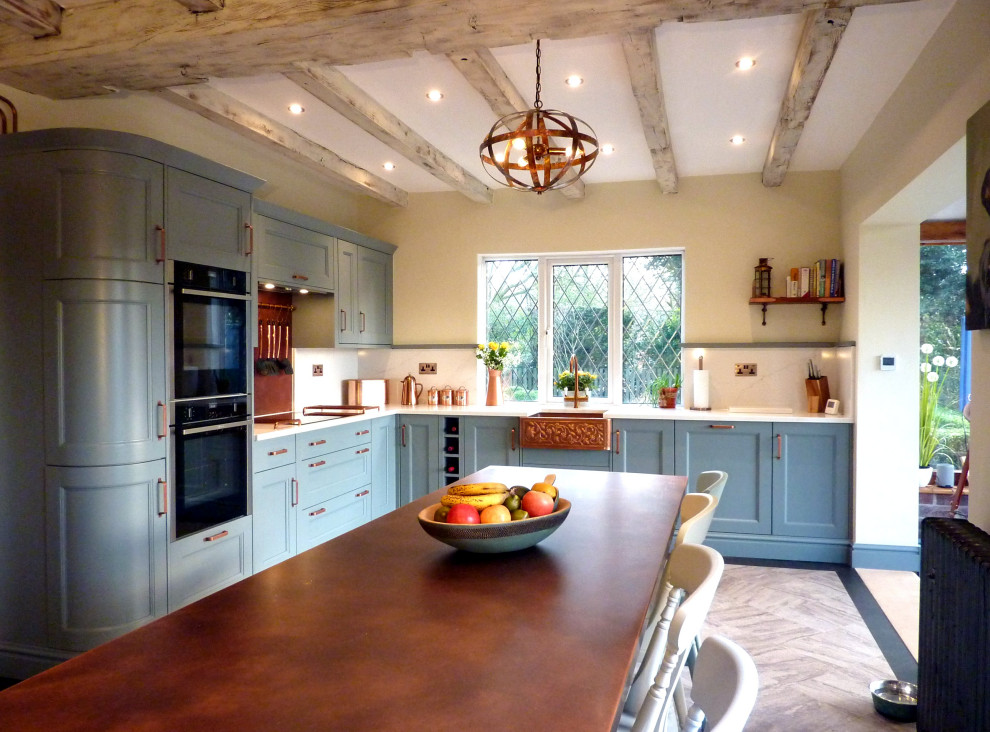 This is an example of a large galley eat-in kitchen in West Midlands with a farmhouse sink, beaded inset cabinets, blue cabinets, quartzite benchtops, orange splashback, black appliances, vinyl floors, with island, grey floor, white benchtop and exposed beam.