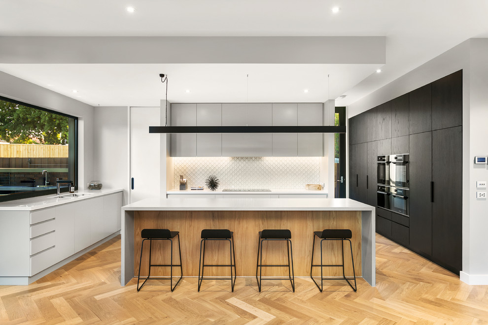 Contemporary u-shaped kitchen in Melbourne with a double-bowl sink, flat-panel cabinets, white splashback, panelled appliances, medium hardwood floors, with island, brown floor and white benchtop.