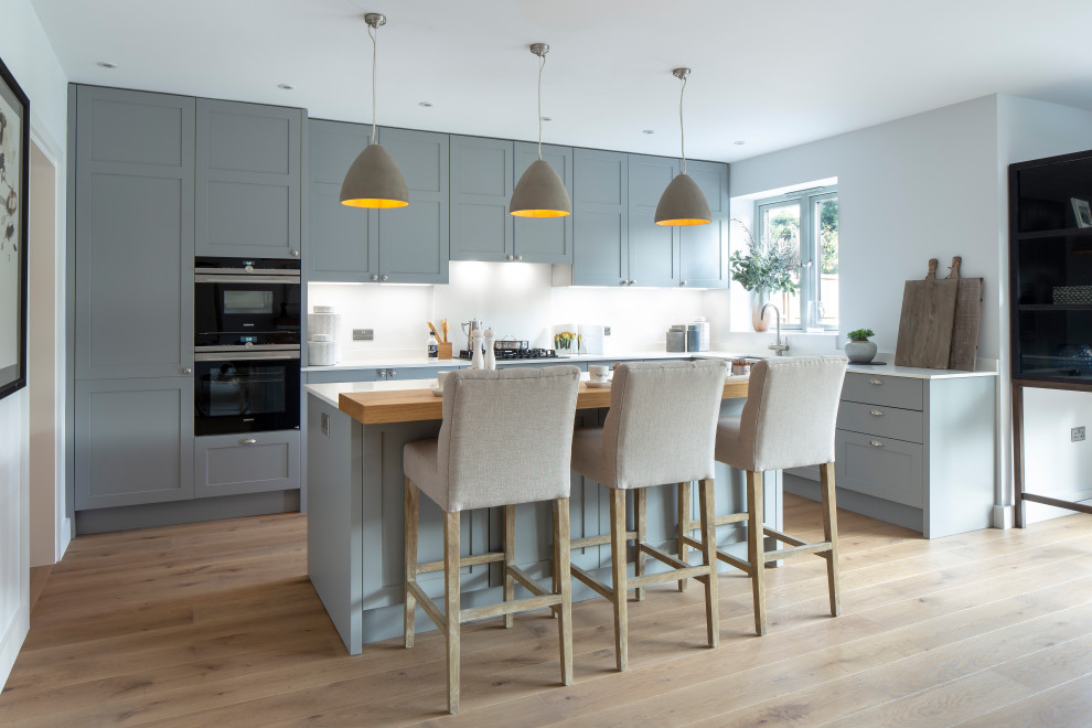 Photo of a traditional l-shaped kitchen in Oxfordshire with a submerged sink, shaker cabinets, grey cabinets, white splashback, stone slab splashback, black appliances, light hardwood flooring, an island, beige floors and white worktops.