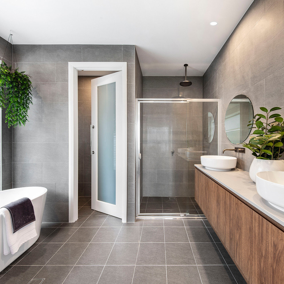 Photo of a mid-sized contemporary master bathroom in Wollongong with furniture-like cabinets, medium wood cabinets, a freestanding tub, a corner shower, a one-piece toilet, gray tile, stone tile, grey walls, cement tiles, a console sink, engineered quartz benchtops, grey floor, a sliding shower screen and white benchtops.