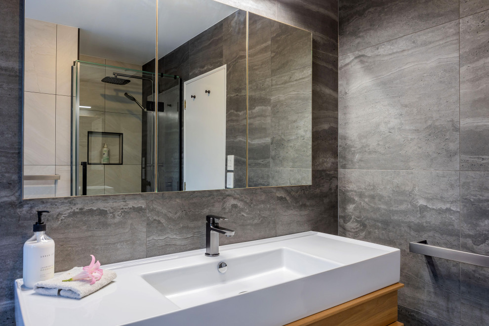 Photo of a small modern ensuite bathroom in Auckland with light wood cabinets, a freestanding bath, an alcove shower, grey tiles, ceramic tiles, porcelain flooring, an integrated sink, solid surface worktops, white floors, a hinged door, white worktops, a single sink and a floating vanity unit.