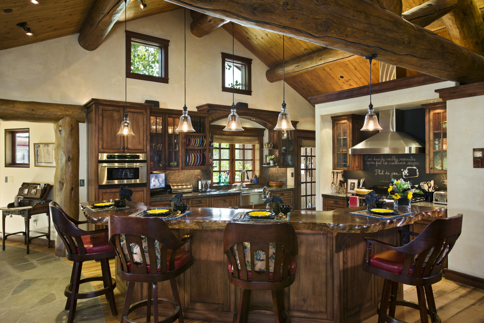 Example of a large mountain style l-shaped medium tone wood floor eat-in kitchen design with a double-bowl sink, glass-front cabinets, dark wood cabinets, wood countertops, stone slab backsplash and an island