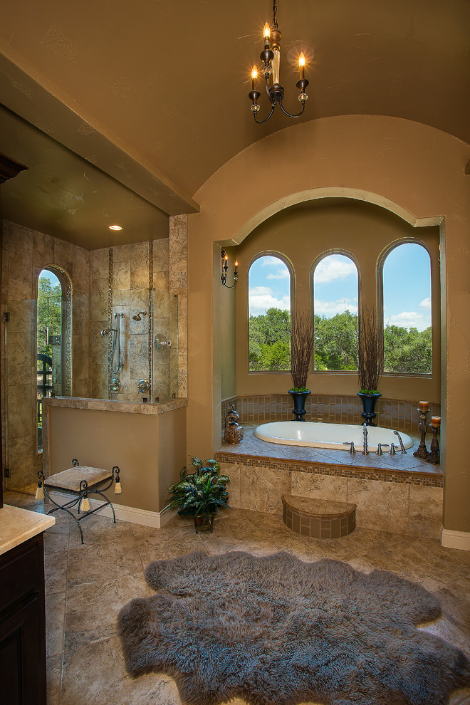 Photo of a large mediterranean master bathroom in Austin with raised-panel cabinets, dark wood cabinets, a drop-in tub, a corner shower, beige tile, stone tile, beige walls, porcelain floors, an undermount sink, marble benchtops, beige floor and a hinged shower door.