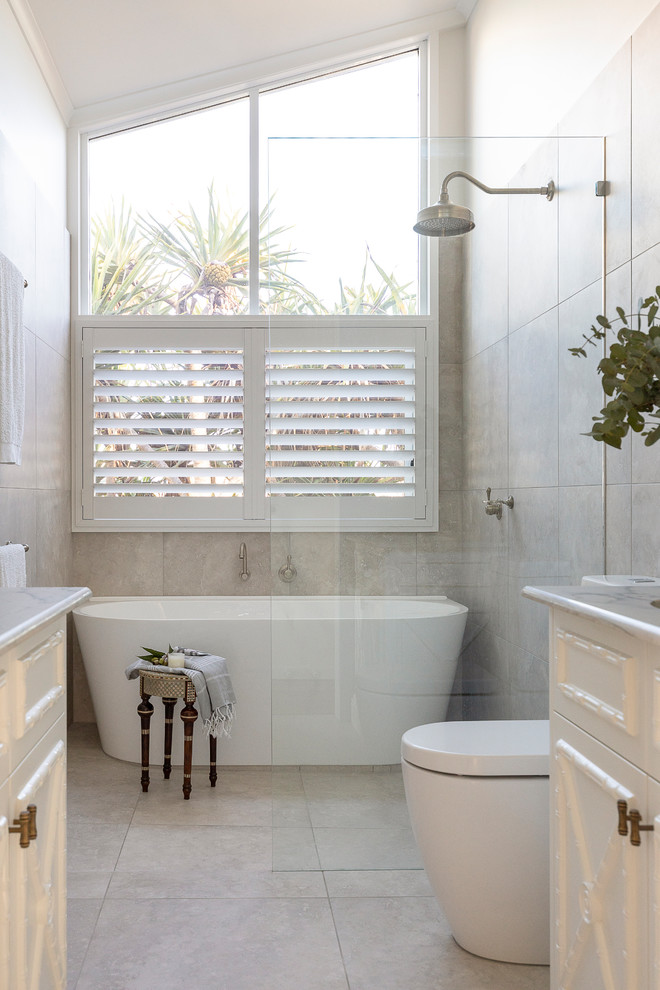 Photo of a mid-sized tropical master bathroom in Sunshine Coast with shaker cabinets, white cabinets, a freestanding tub, an open shower, a wall-mount toilet, beige tile, ceramic tile, ceramic floors, an undermount sink, granite benchtops, beige floor, an open shower and multi-coloured benchtops.