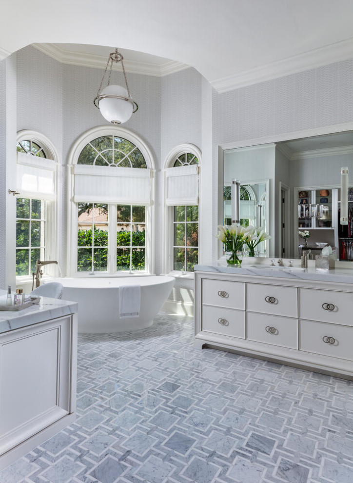 This is an example of an expansive transitional master bathroom in Miami with flat-panel cabinets, white cabinets, a freestanding tub, a curbless shower, a two-piece toilet, white tile, porcelain tile, grey walls, marble floors, an undermount sink, marble benchtops, grey floor, a hinged shower door, white benchtops, a single vanity, a built-in vanity and wallpaper.
