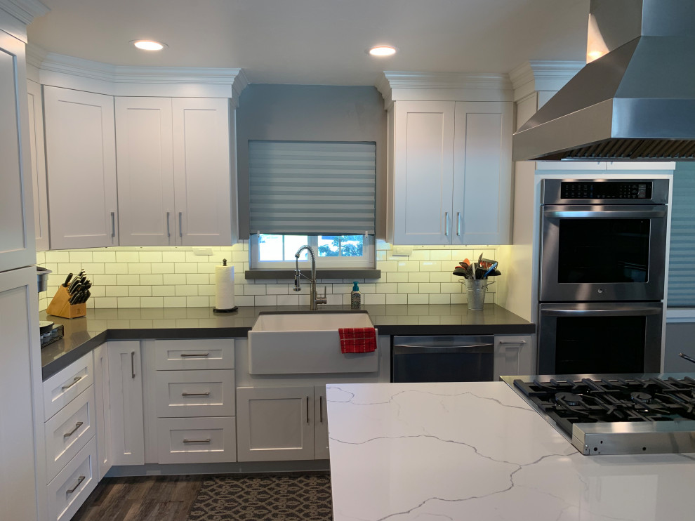 Photo of a mid-sized contemporary l-shaped eat-in kitchen in Sacramento with a farmhouse sink, shaker cabinets, white cabinets, quartz benchtops, white splashback, ceramic splashback, stainless steel appliances, vinyl floors, with island, brown floor and white benchtop.