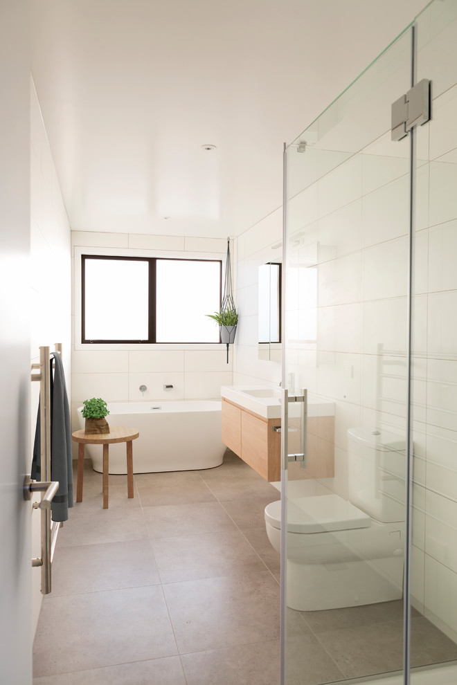 This is an example of a contemporary master bathroom in Auckland with flat-panel cabinets, light wood cabinets, a freestanding tub, a corner shower, a one-piece toilet, white tile, white walls, an integrated sink, beige floor, a hinged shower door and white benchtops.