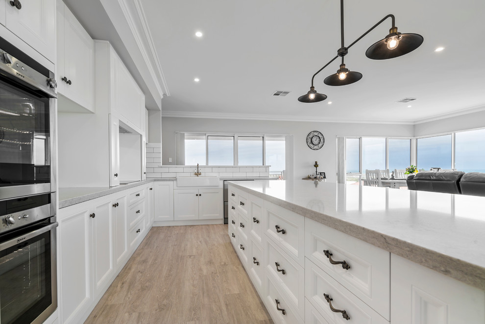 Photo of a mid-sized transitional galley eat-in kitchen in Perth with a farmhouse sink, recessed-panel cabinets, white cabinets, solid surface benchtops, white splashback, ceramic splashback, stainless steel appliances, vinyl floors and with island.