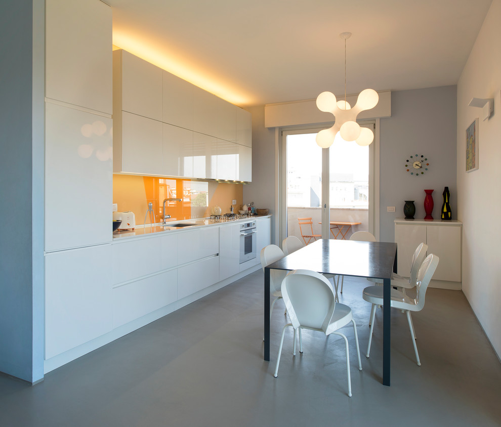 Contemporary single-wall eat-in kitchen in Milan with an undermount sink, flat-panel cabinets, white cabinets, orange splashback, glass sheet splashback, panelled appliances, concrete floors, no island, grey floor and white benchtop.