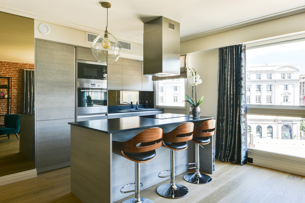 This is an example of a small contemporary galley open plan kitchen in Nice with mirror splashback, stainless steel appliances, light hardwood floors, with island, beige floor, black benchtop, an undermount sink, beaded inset cabinets, light wood cabinets and marble benchtops.